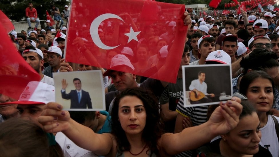 Žena na mitingu okružena crvenim turskim zastavama drži fotografije Selahatina Demirtasa