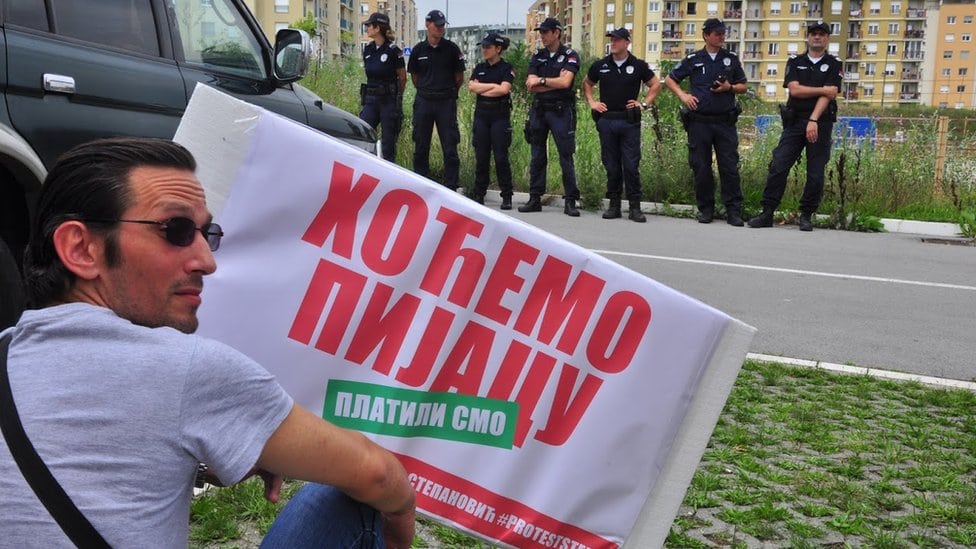Jedan od demonstranata drži transparent na kome piše "Hoćemo pijacu", dok se sedam policajaca vidi u pozadini