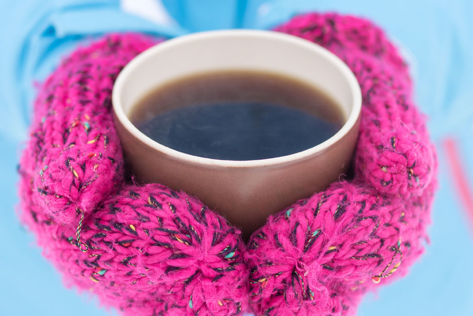 A person wearing mittens clasping a hot drink