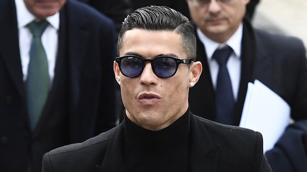Ronaldo in close-up, wearing black clothes, with slick combed-back hair and sunglasses, flanked by men in suits