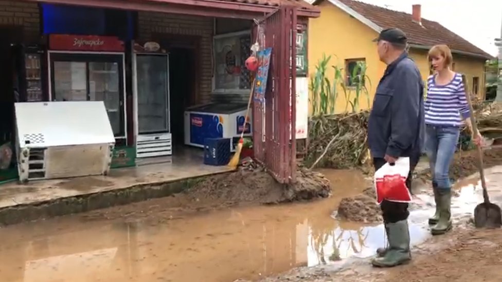 Žena sa lopatom i čovek u gumenim čizmama gledaju u potopljenu seosku prodavnicu.