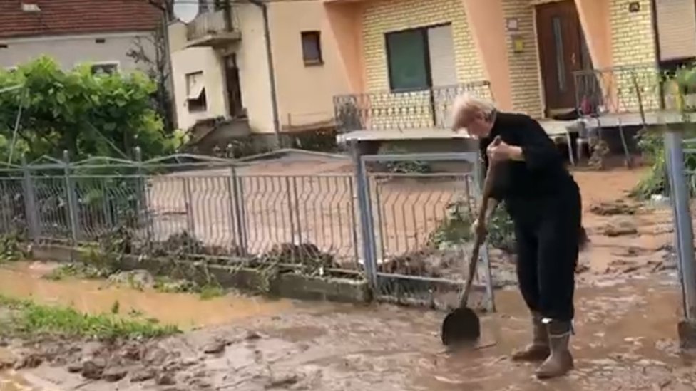 Žena lopatom čisti blato od poplava ispred kuće.