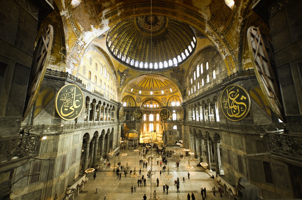Interior of Hagia Sophia captured with fish-eye lens, warm light and unrecognizable people walking about.