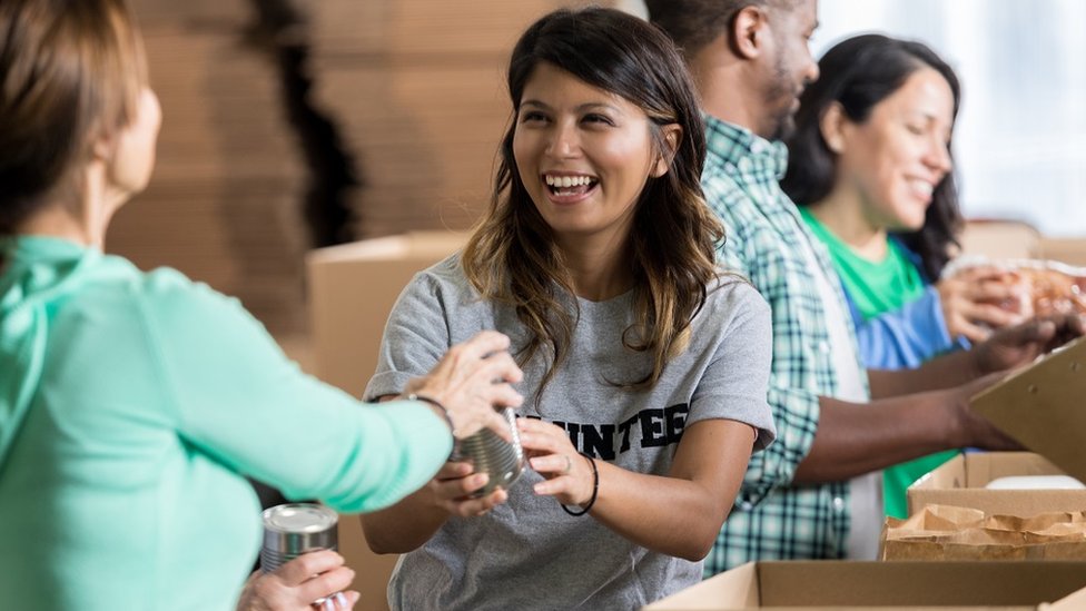 Cheerful mid adult female volunteer accepts a canned food donation during community food drive. She is wearing a gray volunteer t-shirt. Her friends are volunteering in the background.