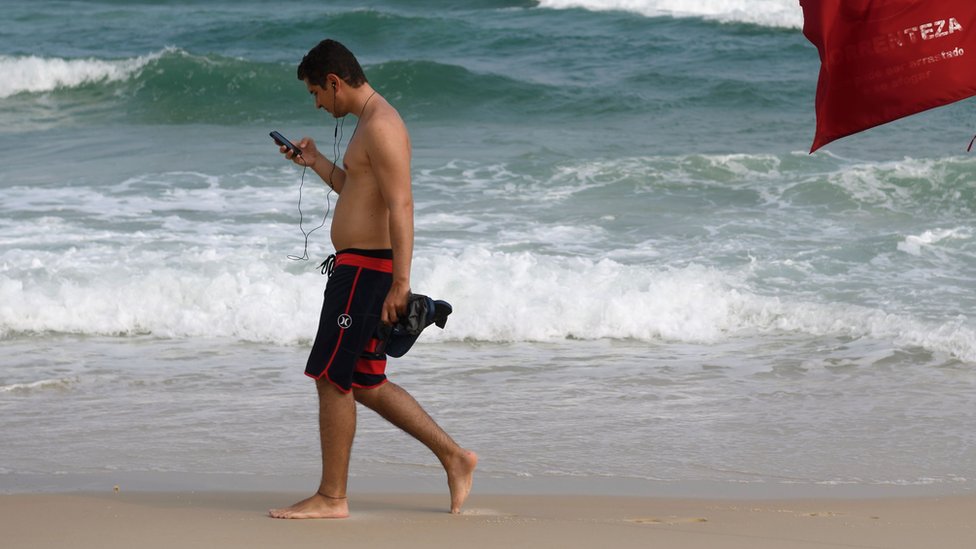 Bez mejlova u slobodno vreme 2 man checking phone while walking down the beach in Brazil
