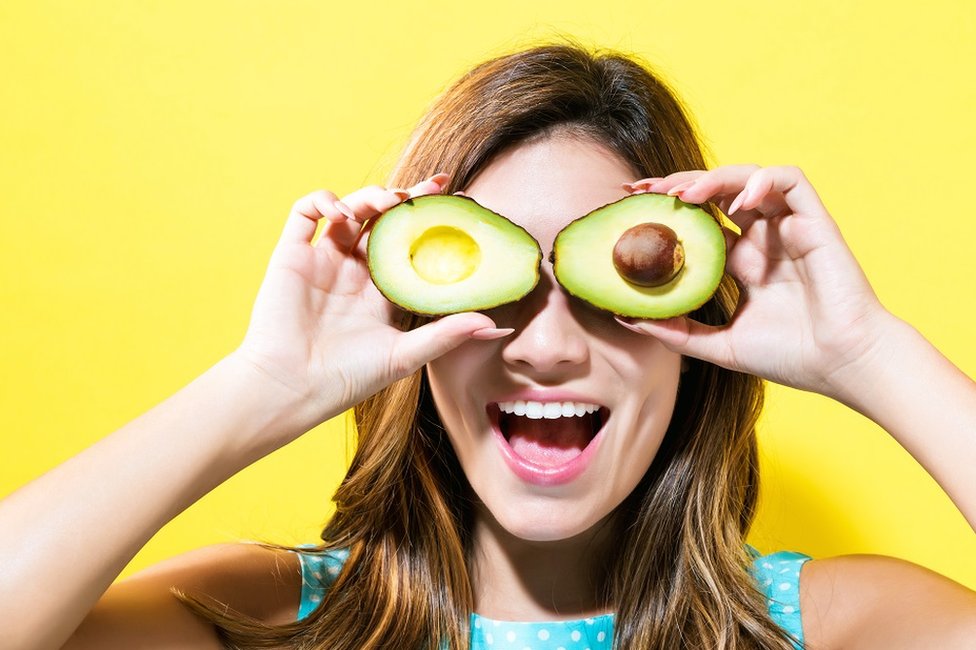 Happy young woman holding avocado halves - bright yellow background