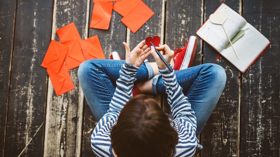 Želite li da poboljšate znanje engleskog? Evo kako da pišete dobro na „najagresivnije nepravilnom jeziku na svetu“ 1 A woman, sitting cross-legged on the floor, writing a note on a heart-shaped paper
