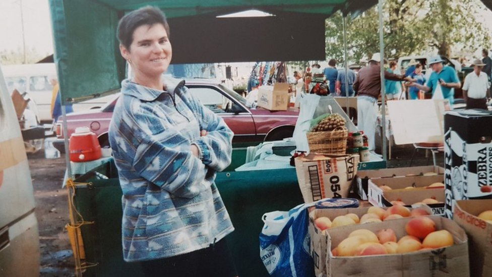 A woman stands at a market with her arms crossed, smiling