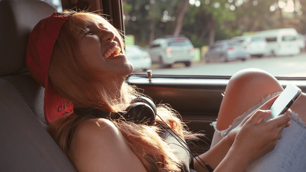 A woman, sitting in a car, laughing with a phone in her hand