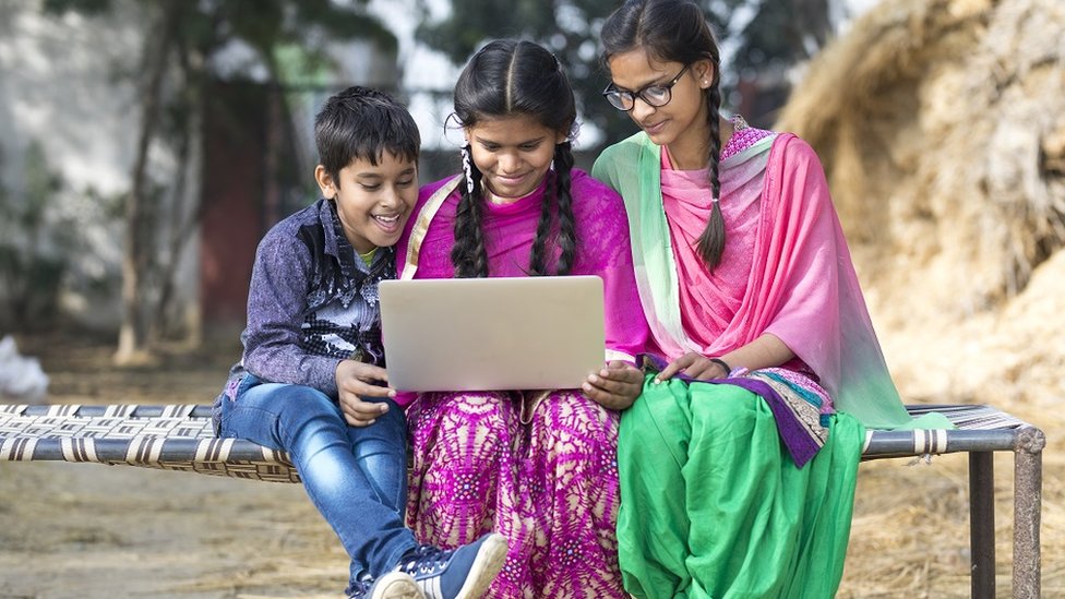 Two teenage girls and a boy, dressed in traditional Indian attire, using a laptop while sitting outdoors