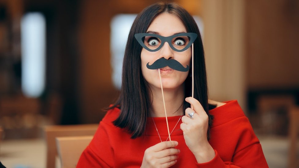 Funny woman holding a paper party mask over her eyes and a fake paper moustache over her mouth
