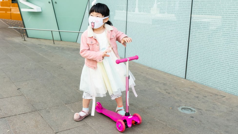 Small Chinese girl, wearing a mask, on a pink scooter going out and about