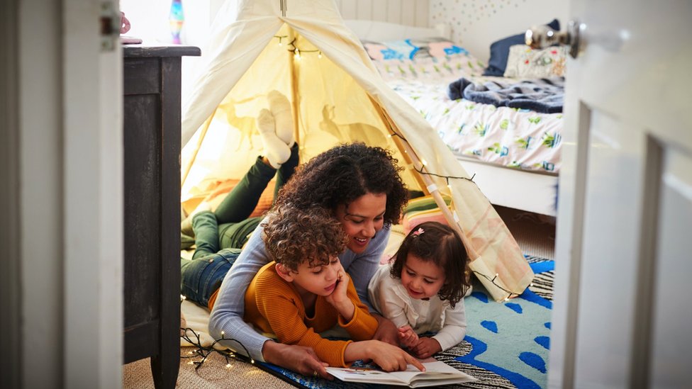 Letovanje kod kuće tokom pandemije: Kako uživati u odmoru bez putovanja 7 A woman reading a book to her two children under a tent in the bedroom