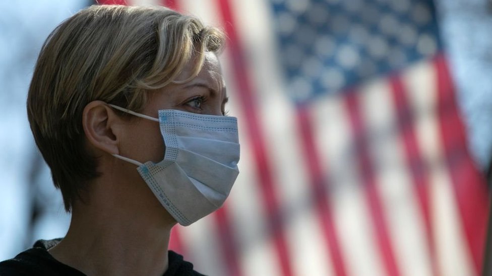 A woman wearing a mask stands in front of a US flag