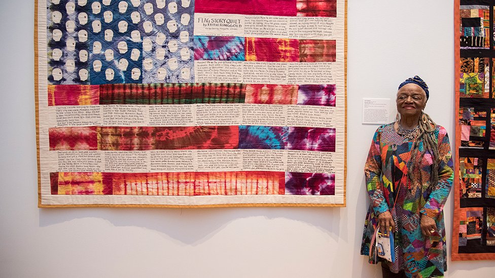 Artist Faith Ringgold standing next to one of her quilted artworks titled "Flag Story Quilt"