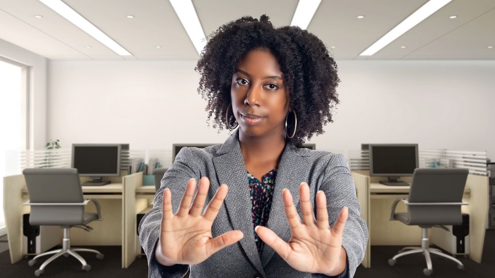 A woman in her office stands with her hands out in front of her indicating "no"