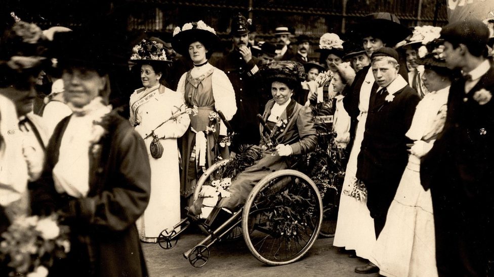 A black and white photo of Rosa at a rally surrounded by her fellow suffragettes. She wears a hat and a long dress.