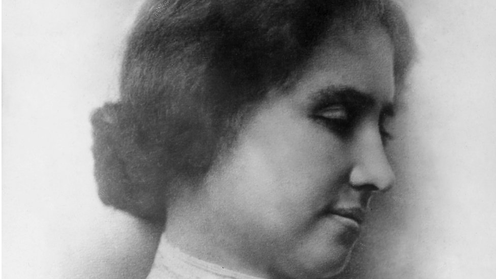 A black and white photo of Helen Keller's side profile. She has brown hair in a low bun and her eyes are downcast