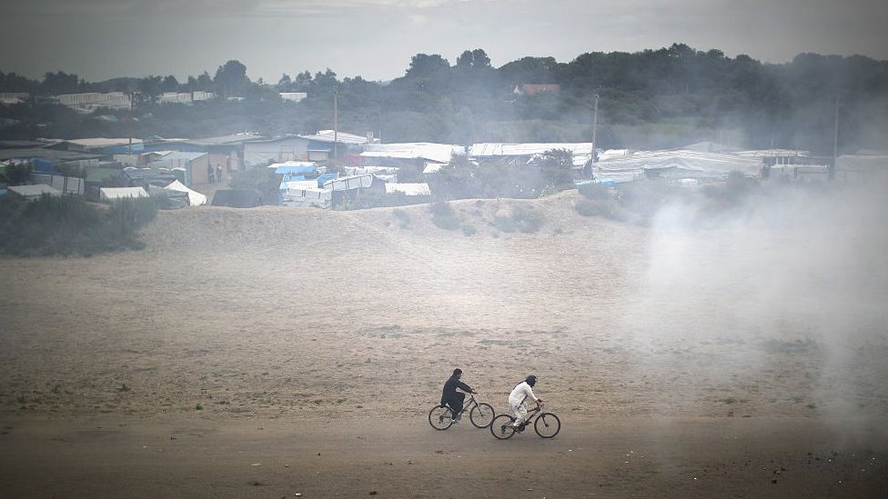 Migrants cycle through smoke from a fire burning rubbish at the Jungle migrant camp on September 6, 2016 in Calais, France.