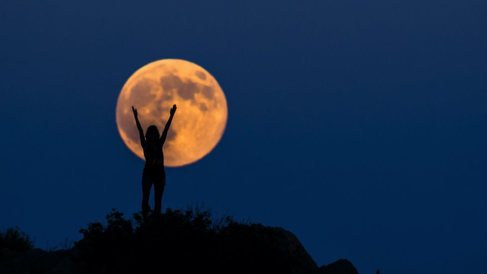 An orange full moon, with a woman in front of it, arms in the air.