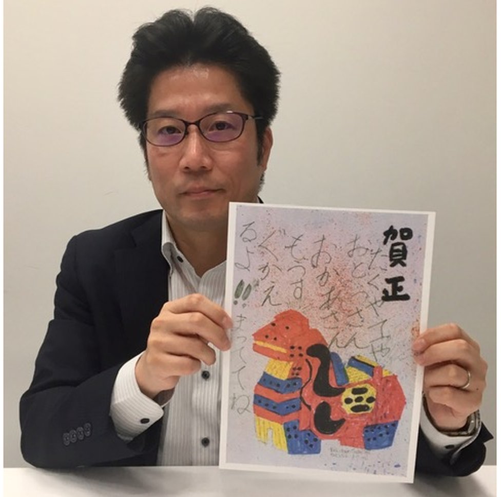 Takuya Yokota, in a black suit, holds up a copy of a postcard featuring a colourful animal