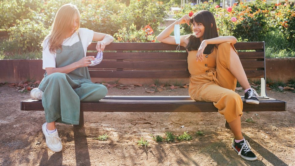 Two girls sitting on a park bench, holding face masks in their hands and socially distancing