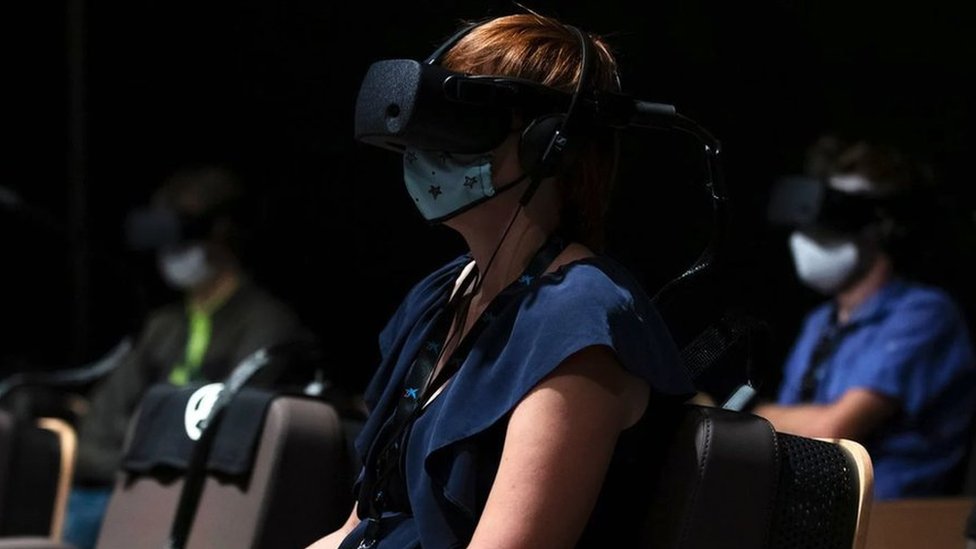 A woman at a film screening room, wearing virtual reality goggles and a protective face mask over her nose and mouth.