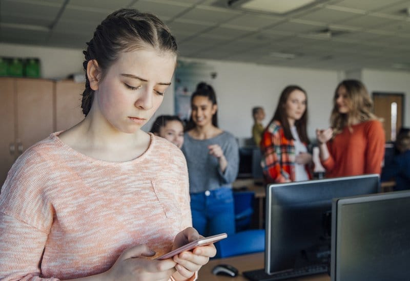 Društvene mreže: Ko je maskirani osvetnik sa TikToka 2 Stock image of a teenage girl looking at a bullying comment on her phone, in a classroom surrounded by other teenagers.