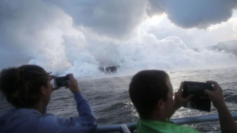 People take photos on a tour boat as steam plumes rise while lava enters the Pacific Ocean, after flowing to the water from a Kilauea volcano fissure, on Hawaii's Big Island on May 20, 2018 near Pahoa, Hawaii.