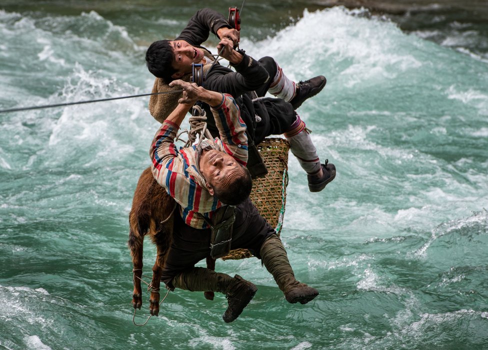Dvojica muškaraca i životinja se prevoze žičarom iznad nabujale reke