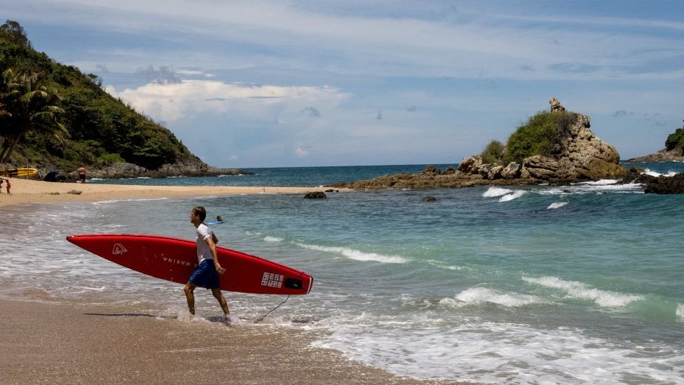 Korona virus i turizam: Pandemija preti da pomrsi planove za početak turističke sezone u Tajlandu 1 A man carrying his paddle board out of the water on a beach in Phuket