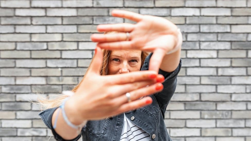 Close-up of woman against grey brick wall making finger frame sign