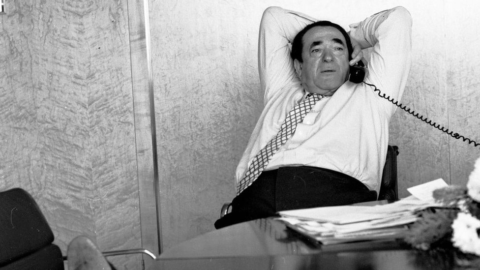 Robert Maxwell leans back in his chair as he talks on an office phone with his feet resting on another chair