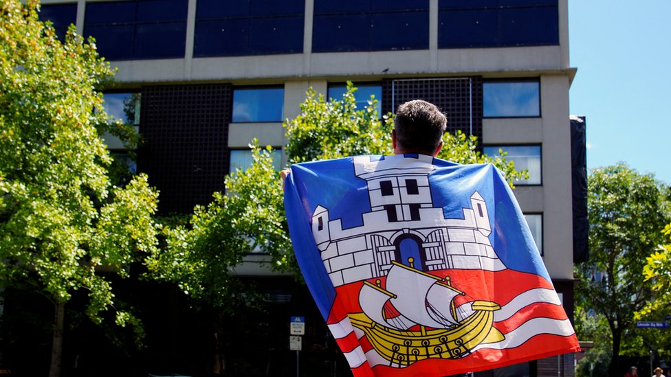 Novak Đoković, vakcine i Australija: Zašto je srpski teniser ostao bez vize 2 A fan draped in the Serbian flag looks up at the Park Hotel in Melbourne where Novak Djokovic has reportedly been detained