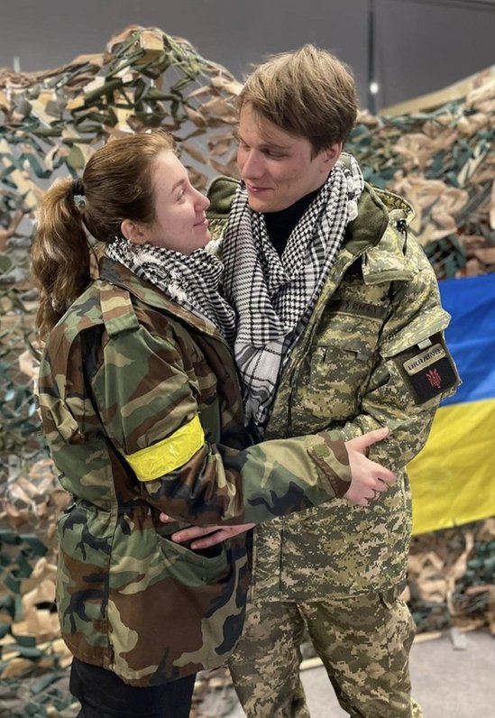 Yaryna Arieva and Svyatoslav Fursin hugging in military uniforms with a Ukrainian flag behind them