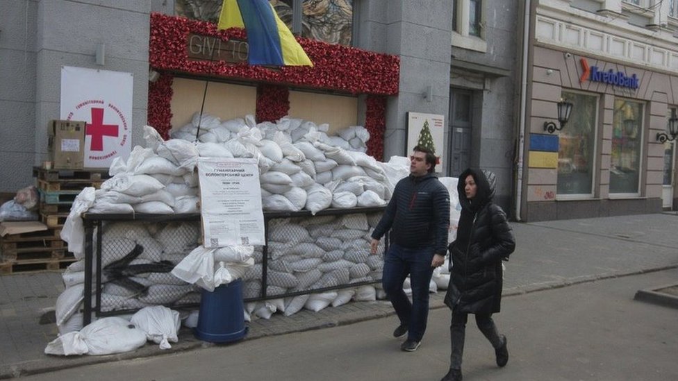 Two people walking down a street past a building protected by sandbags