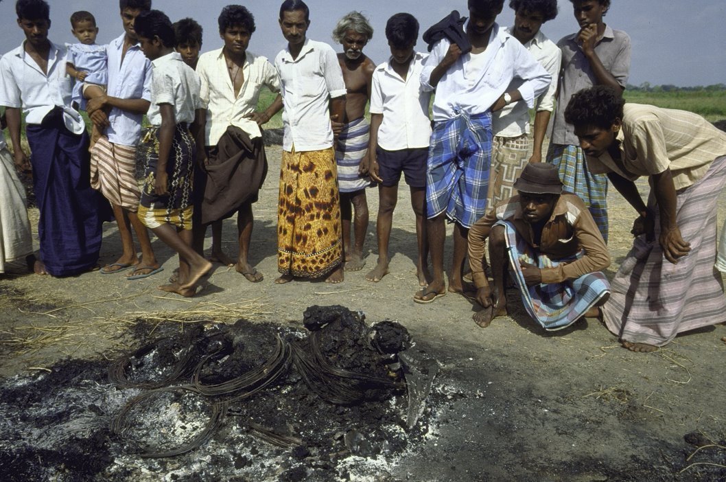 Burnt tyres lying on the ground with unidentifiable remains among them, while a group of men look on
