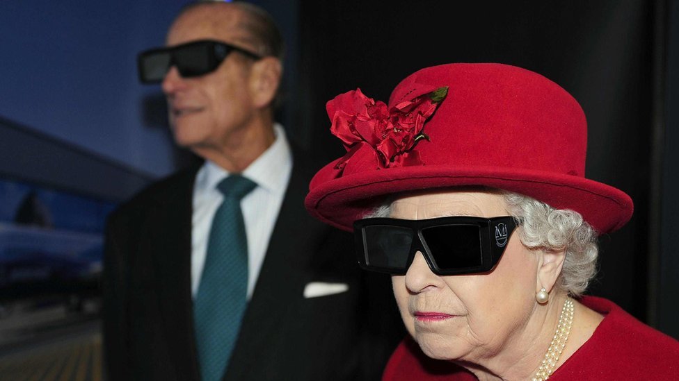Prince Philip and the Queen in red hat and 3d dark glasses