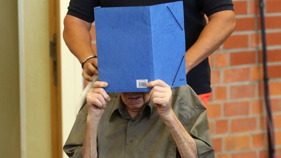 Former Nazi concentration camp guard Josef S (R) hides his face with a folder as he arrives on June 28, 2022 at a gym used as a makeshift courtroom in Brandenburg an der Havel, eastern Germany, where his verdict was spoken.