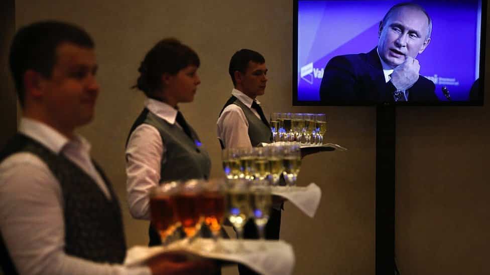 Vladimir Putin i korona virus: Kako Rusija troši čitavo bogatstvo na zaštitu predsednika od kovida 5 Waiters hold trays of drinks while Putin appears on a TV screen