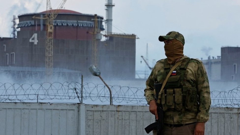 A masked man with an assault rifle stands in front of the nuclear power plant