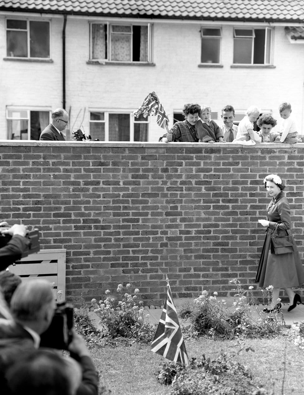 Kraljica Elizabeta Druga: Život u fotografijama 16 A Union Jack planted in the garden caught the Queen's attention, but she is the object of the neighbours' gaze. They watched as she arrived to visit the home of Mr and Mrs Eddie Hammond at Crawley New Town, Sussex after opening Gatwick Airport. 1958