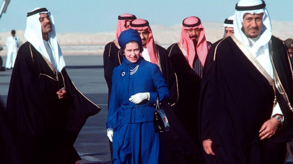 Queen Elizabeth during her state visit to Saudi Arabia, standing among a number of male members of the local delegation, all of whom are wearing traditional dress - the Queen is wearing a blue head cover that matches her body clothing, while the delegates are wearing head coverings in their traditional attire