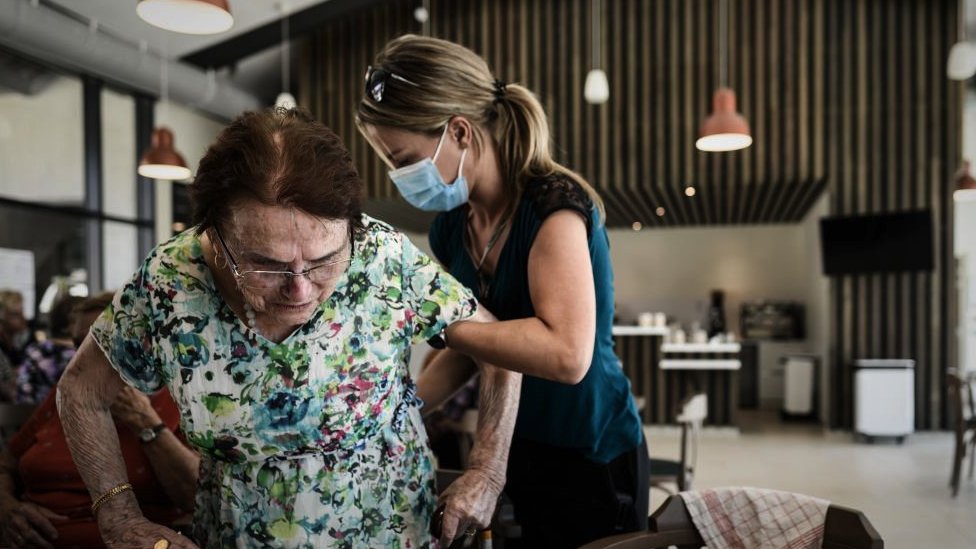 A volunteer helps support a woman standing above a chair at a table during lunch at the Landais Alzheimer site for Alzheimers patients in Dax, France