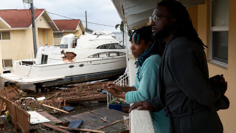 A man and woman stand on their balcony next to a boat that smashed into their apartment complex.