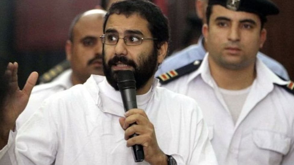 Alaa Abdel Fattah speaks into a microphone as men in uniform stand behind him