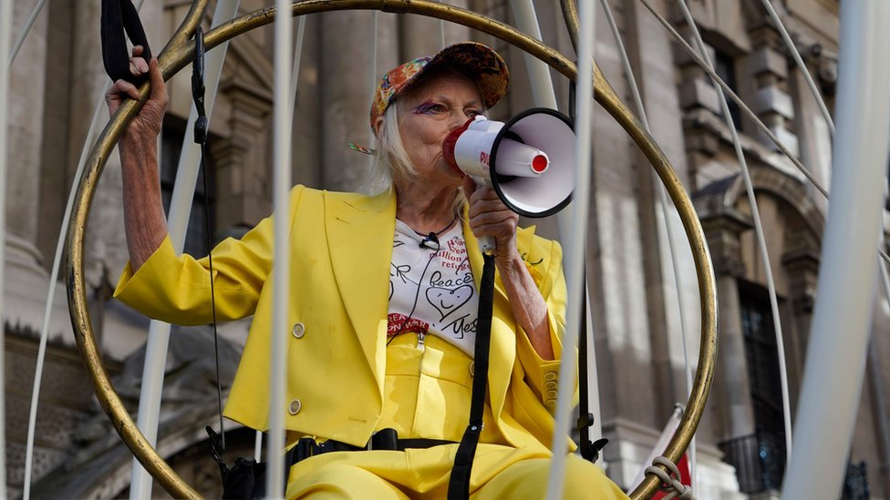Vivienne Westwood speaks into a megaphone while dressed in yellow in a bird cage