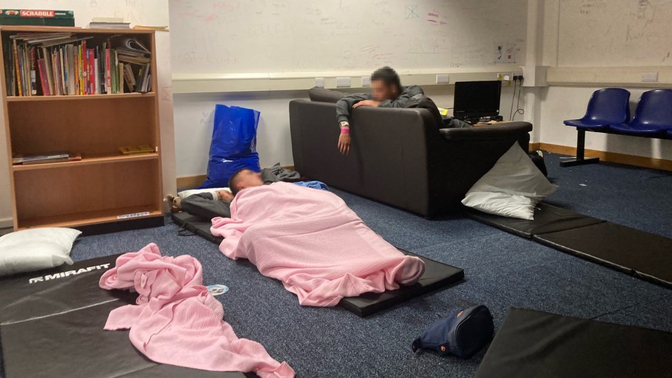 One man lies on the floor under a pink blanket while another sits on a sofa near a bookcase