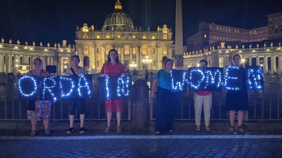 Women hold a sign saying Ordain Women