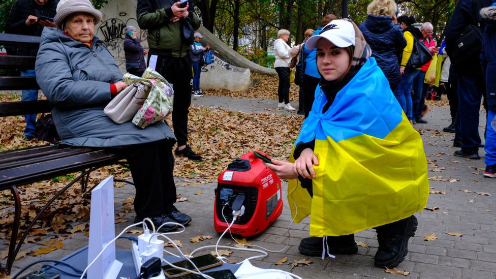 Rusija i Ukrajina: Eksplozije odjekuju Kijevom, gradonačelnik kaže da Rusi ponovo napadaju dronovima 3 Woman draped in Ukrainian flag uses generator to charge phone